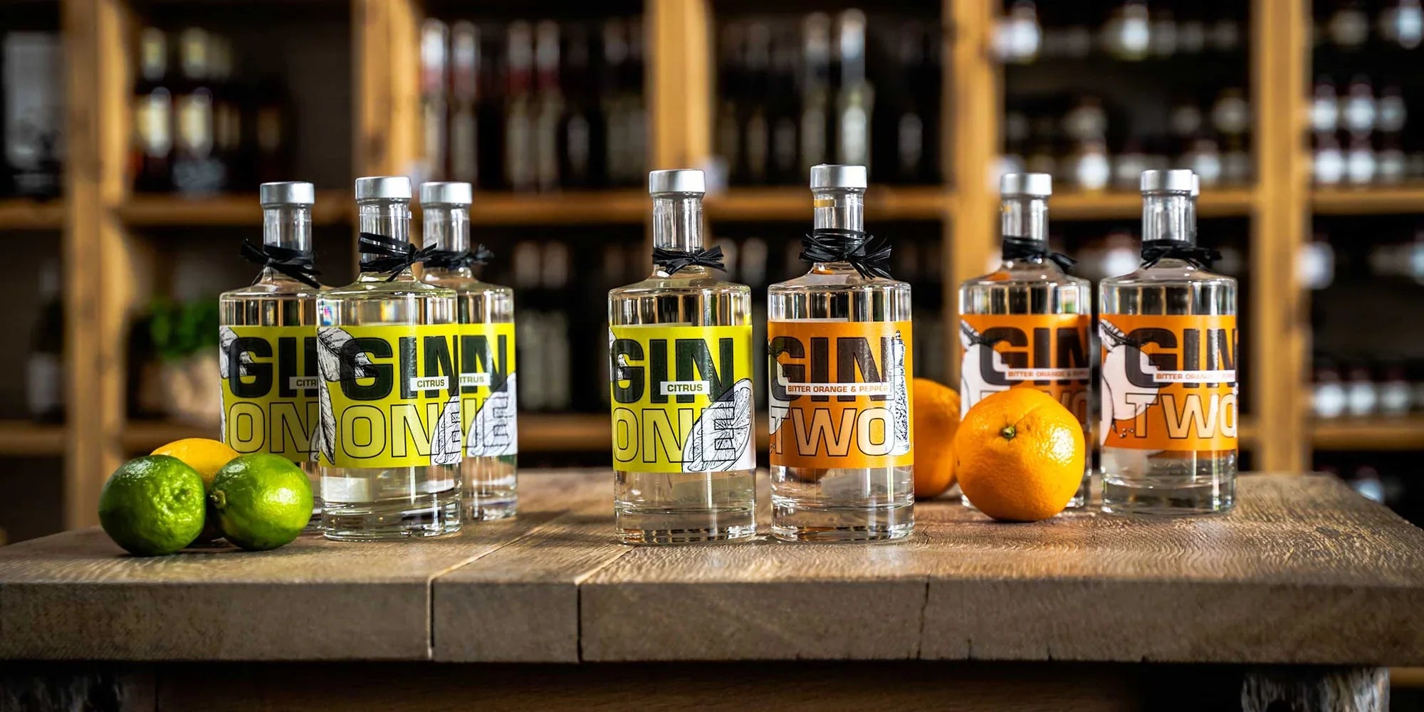 Eine Auswahl an Gin, vielfältig wie Parfüm, mit Orangen und Limetten auf einem Tisch vor einem Weinregal.